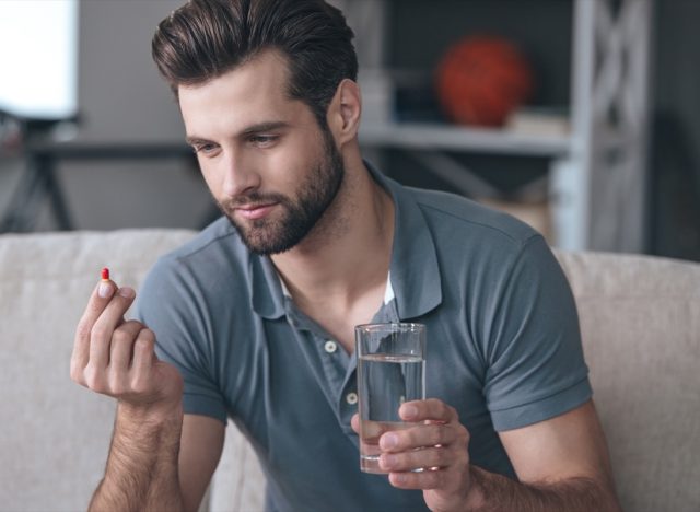 man holding a glass of water and looking at a pill in his hand while sitting on the couch