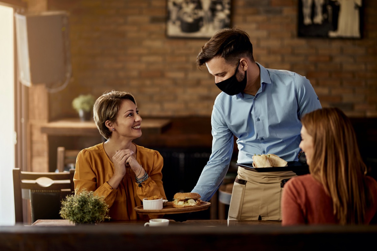 Young waiter wearing protective face mask while serving food to his guests in a restaurant.