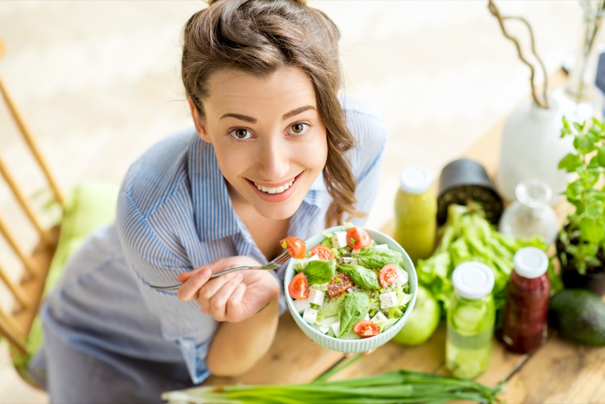 femeie fericită care mănâncă salată sănătoasă așezată pe masă cu ingrediente proaspete verzi în interior