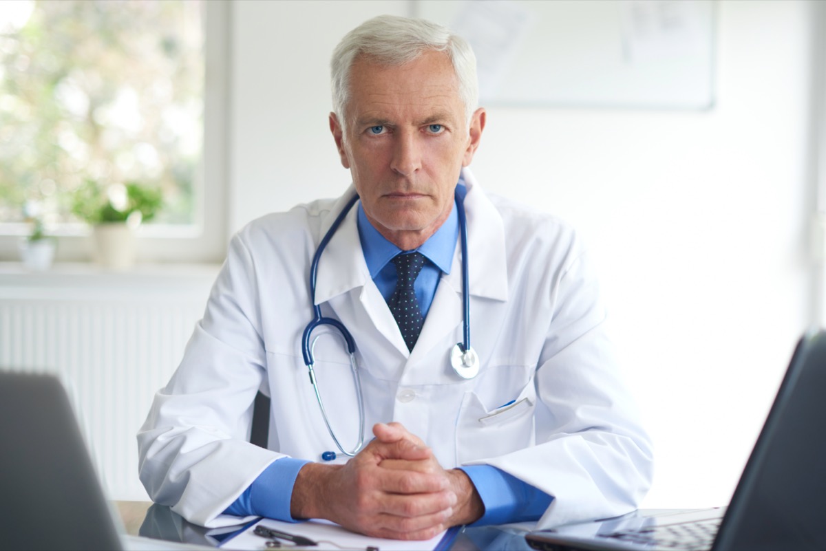 Portrait of senior male doctor sitting at doctor's office and working on laptop.