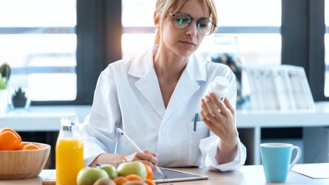 Shot of woman nutritionist doctor writes the medical prescription for a correct diet on a desk with fruits, pills and supplements.