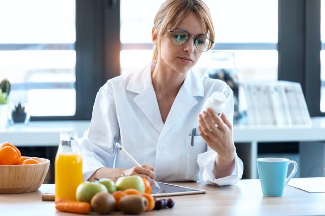 Bir kadın diyetisyen doktorun bir fotoğrafı, meyve, hap ve takviyelerle dolu bir masaya uygun bir diyet reçetesi yazar.