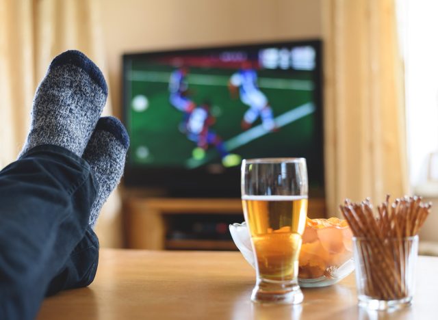 Man watching a sports game on tv while sitting on a couch eating junk snacks and drinking a beer