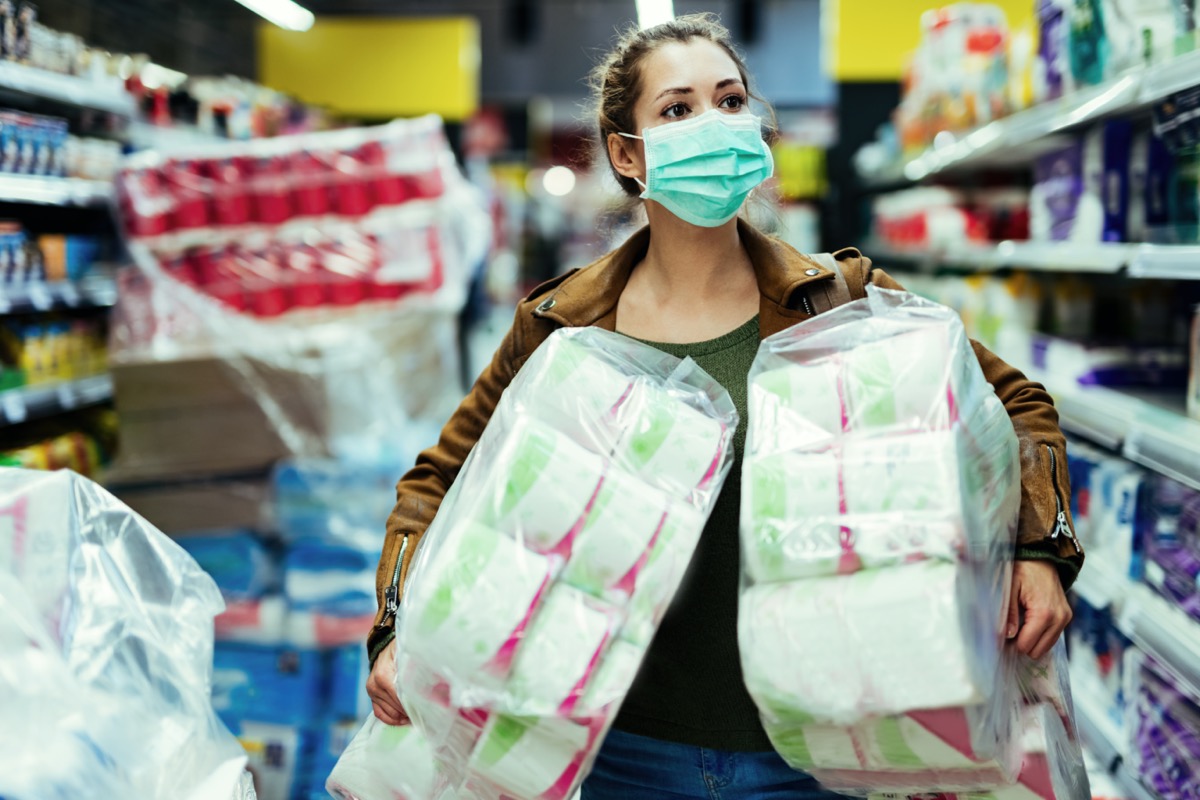 Woman wearing protective face mask while carrying packages of toilet paper and buying in time of virus pandemic.