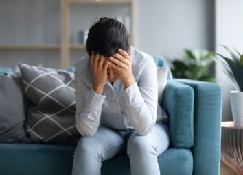 depressed Indian woman holding head in hands, sitting alone on couch at home