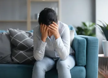 depressed Indian woman holding head in hands, sitting alone on couch at home