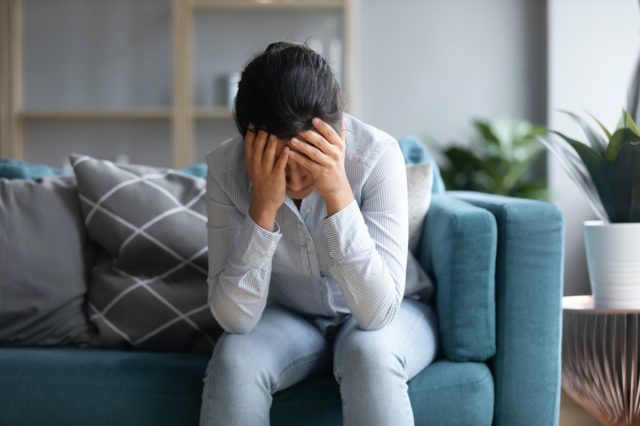 depressed Indian woman holding head in hands, sitting alone on couch at home