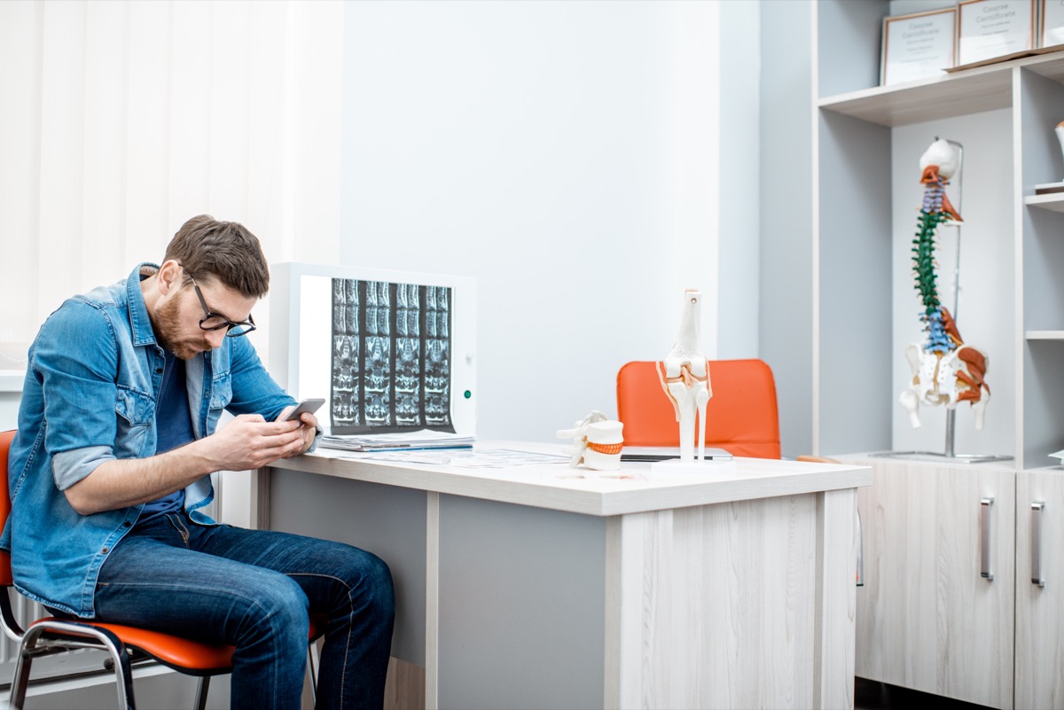 Man using smartphone sitting hunched down having scoliosis in the office of therapist at the clinic