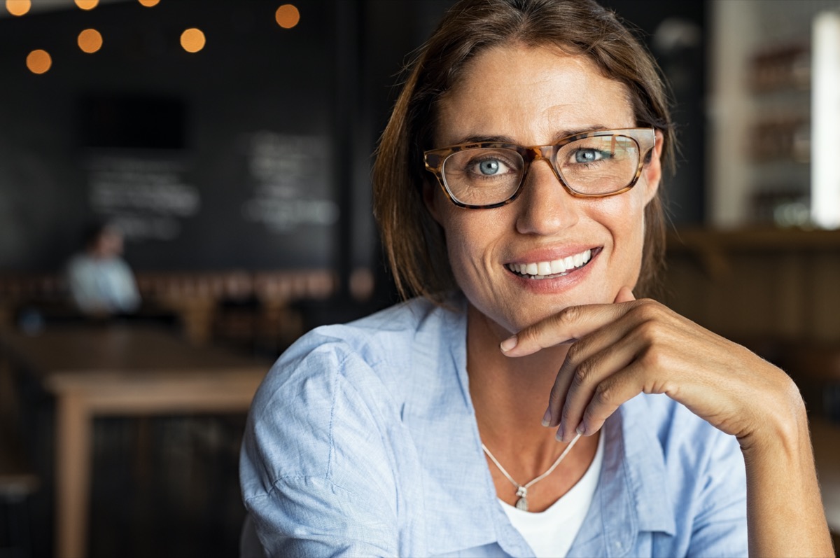 Portrait of happy mature woman wearing eyeglasses and looking at camera. Closeup face of smiling woman sitting in cafeteria with hand on chin. Successful lady in a cafe pub.