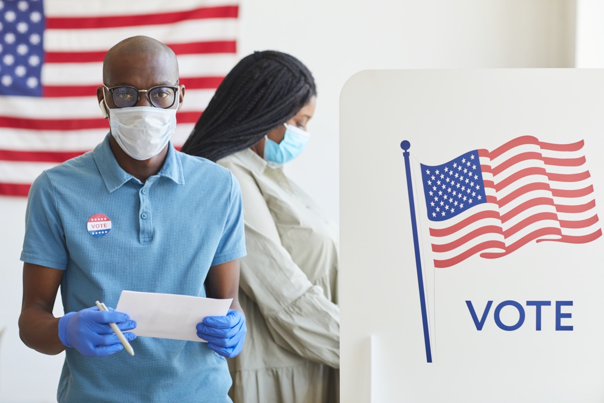 Portrait of a man standing by voting booth decorated with USA flag and looking at camera on post-pandemic election day.