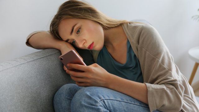 woman with depressed facial expression sitting on grey textile couch holding her phone