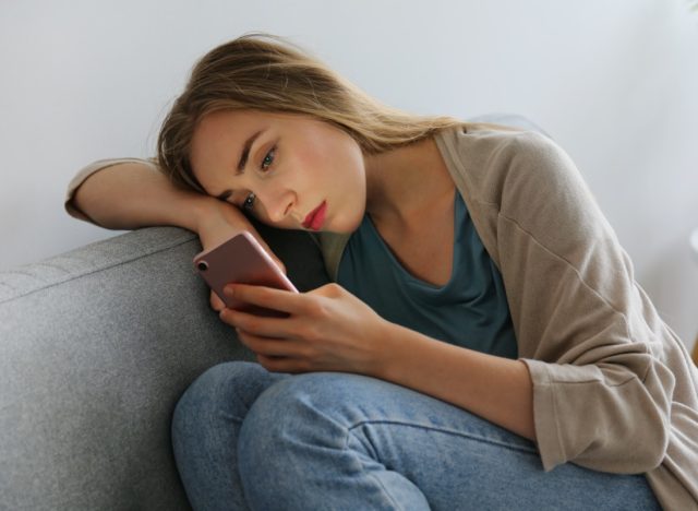 woman with depressed facial expression sitting on grey textile couch holding her phone