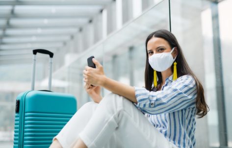 woman with smartphone going on holiday, wearing face masks at the airport