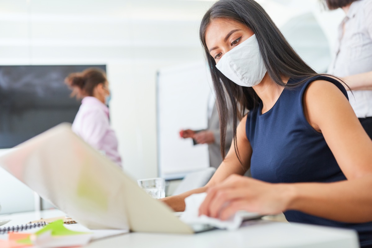 Businesswoman using face mask while disinfecting keyboard in office.