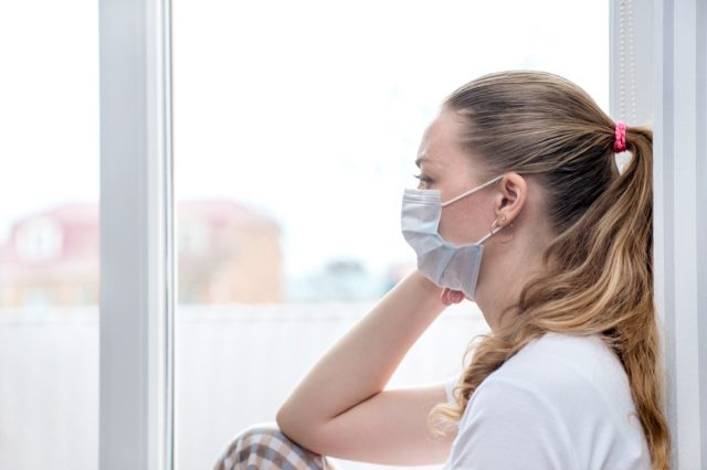 Woman sits at window in a medical mask.