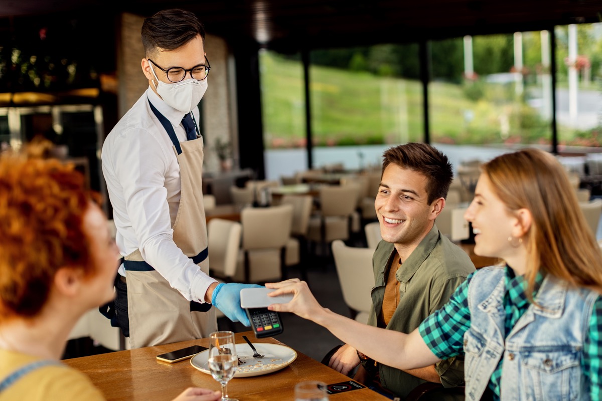 Group of friends paying contactless with mobile phone to a waiter in a cafe.