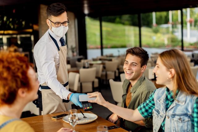 Group of friends paying contactless with mobile phone to a waiter in a cafe.