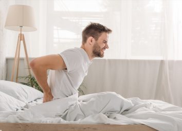 Side view of a frowned young man suffering from pain in loin while sitting on white bedding