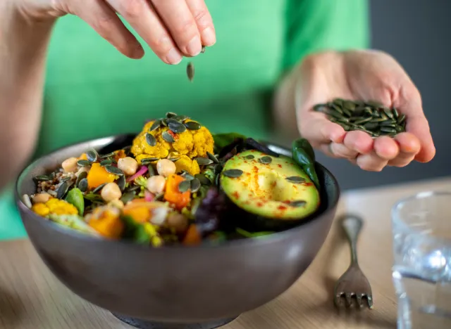 Woman adding pumpkin seeds to vegetable salad bowl and avocado plant meal