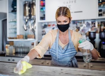 Young woman with face mask working indoors in cafe, disinfecting counter.