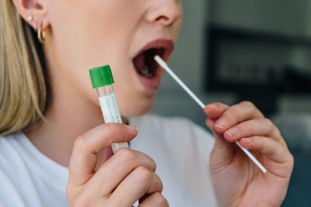 Woman holds a swab into her mouth and holding a medical tube for the coronavirus home test.