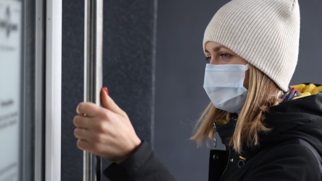 Young woman in protective medical mask and warm clothing opening door to store