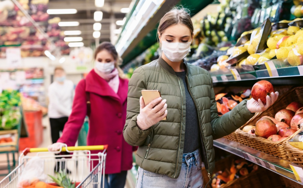 Woman in a disposable face mask is checking a shopping list on a smartphone in a supermarket