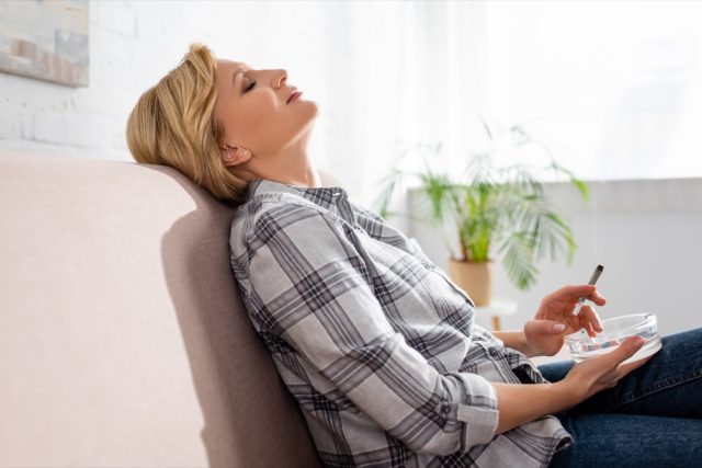 A mature woman with closed eyes sitting on sofa and holding joint with legal marijuana.
