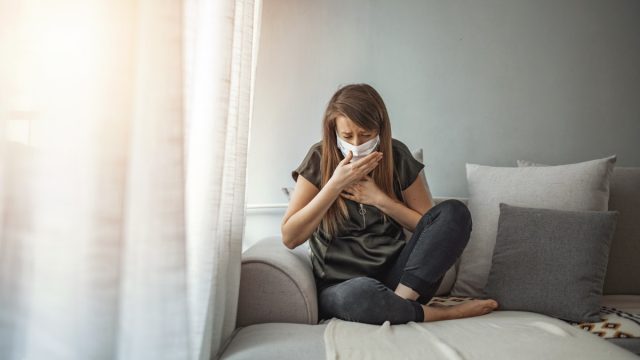 Woman wearing surgical mask on face protective for spreading of disease Covid-19 pandemic.. Girl symptom cough while sitting on Sofa.