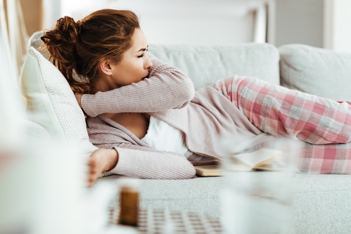 woman coughing into elbow while lying down on sofa in the living room.