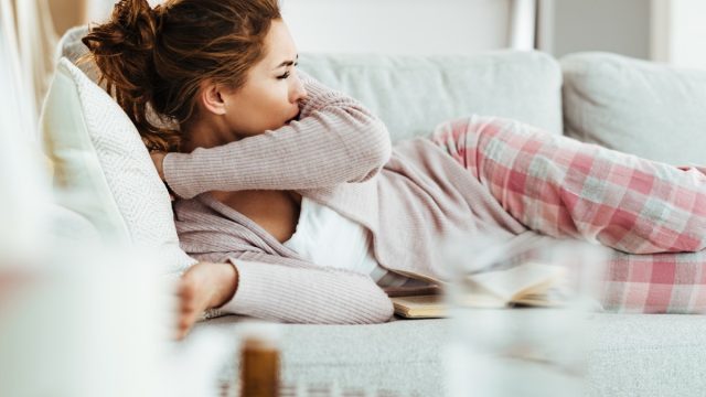 woman coughing into elbow while lying down on sofa in the living room.