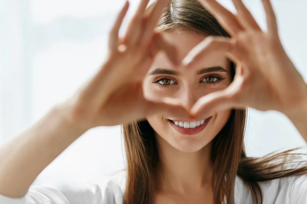 Happy Woman Holding Heart Shaped Hands Near Eyes. Closeup Of Smiling Girl With Healthy Skin Showing Love Sign