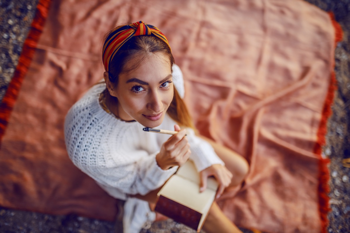 Top view of Caucasian brunette in sweater and headband sitting on blanket outdoors and holding pen and diary in her hands as she looks at the camera.