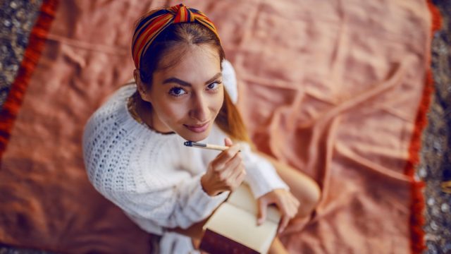 Top view of caucasian brunette in sweater and with headband sitting on blanket outdoors and holding pen and diary in hands while looking at camera.