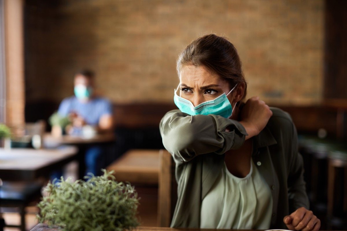 Woman with face mask sneezing into elbow while sitting in a cafe.