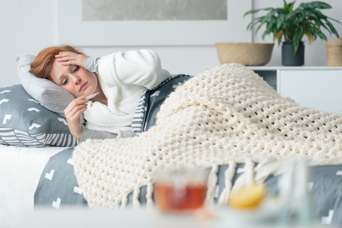 Ill woman lying in bed looking at thermometer suffering from seasonal flu and infectious disease