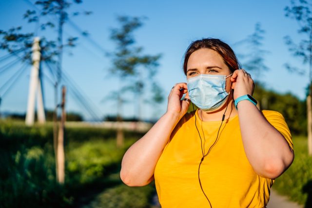 Woman with protective face mask setting music on smart phone and putting earphones before exercise.