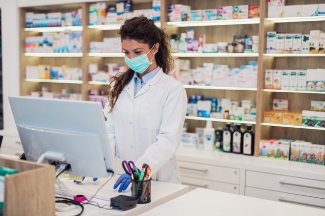 Female pharmacist with protective mask on her face working at pharmacy.