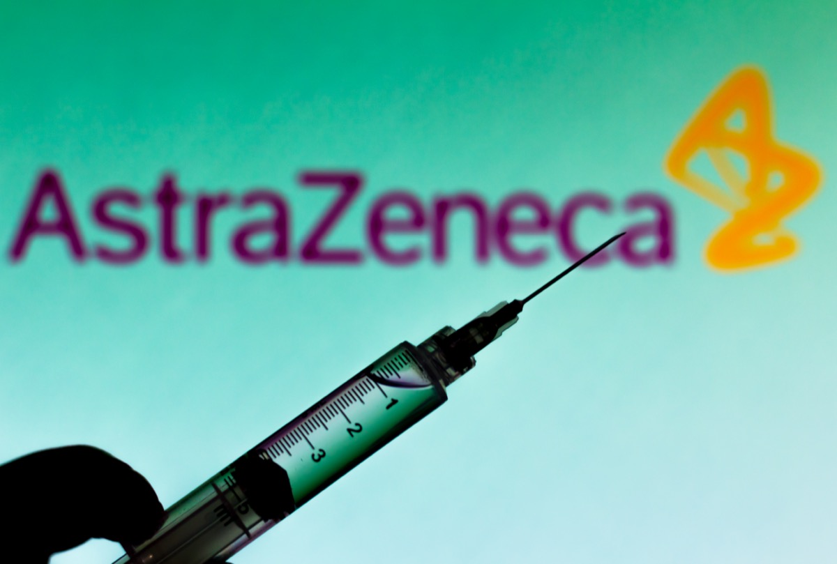 the medical syringe is seen with AstraZeneca company logo displayed on a screen in the background