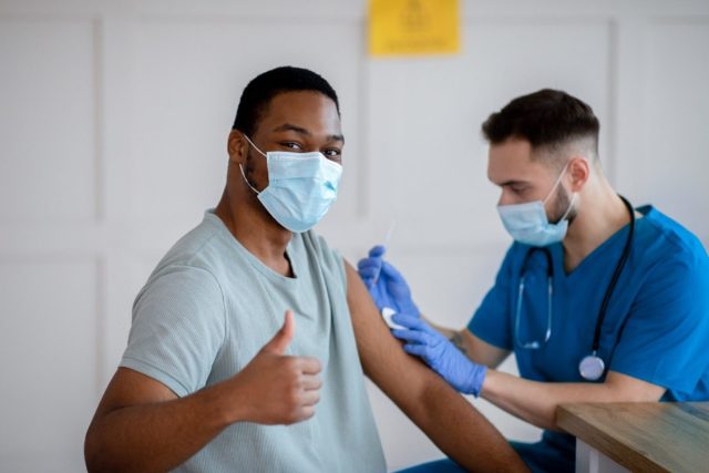 African american man in antiviral mask gesturing thumbs up during coronavirus vaccination, approving covid-19 immunization