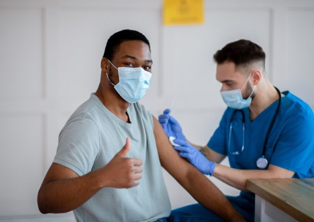 African American man in antiviral mask gesturing thumb up during coronavirus vaccination, approving of covid-19 immunization