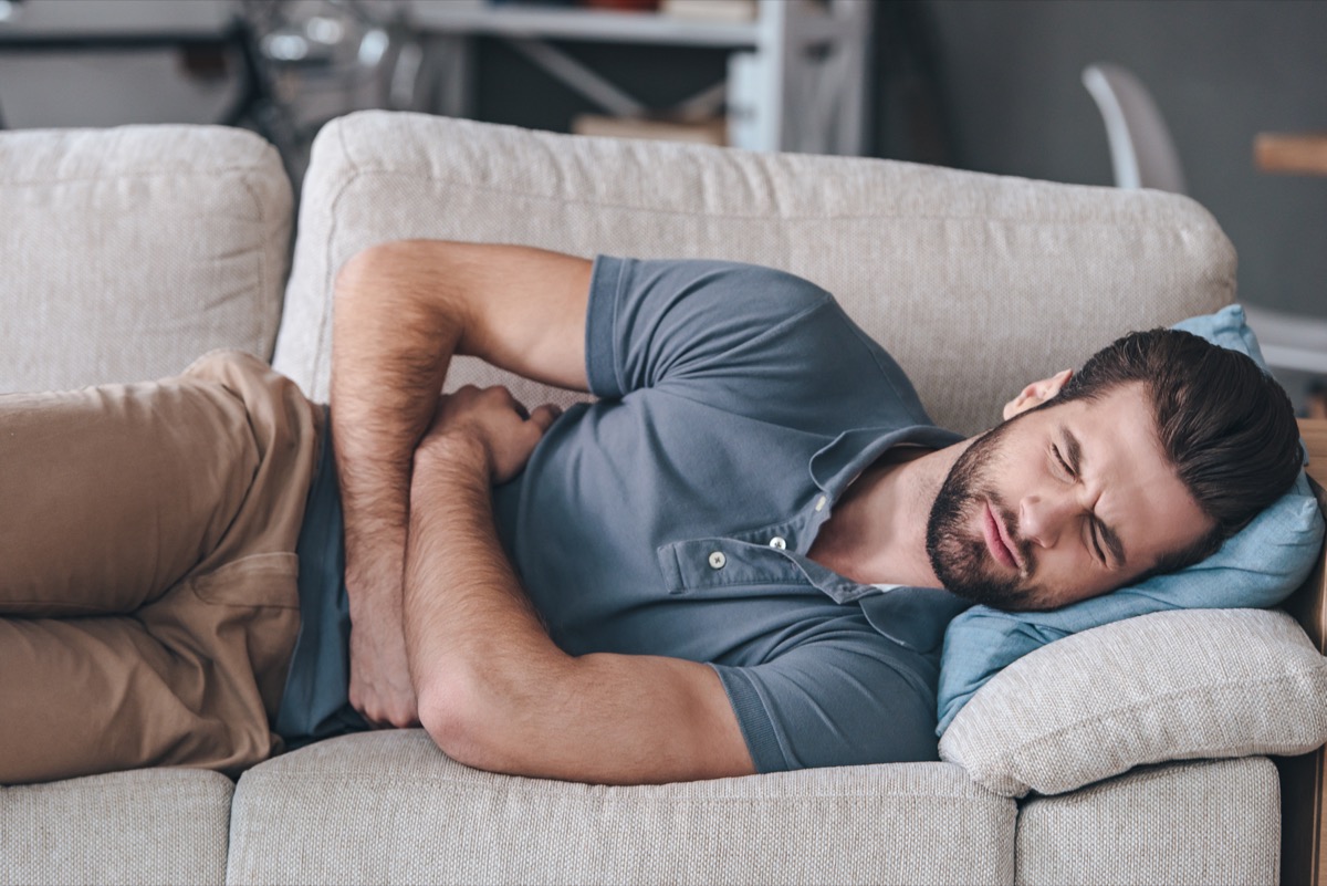 Man hugging his belly and keeping eyes closed while lying on the couch at home