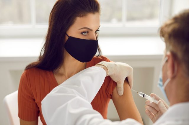 Woman wearing medical protective mask receiving injection in arm vaccination.