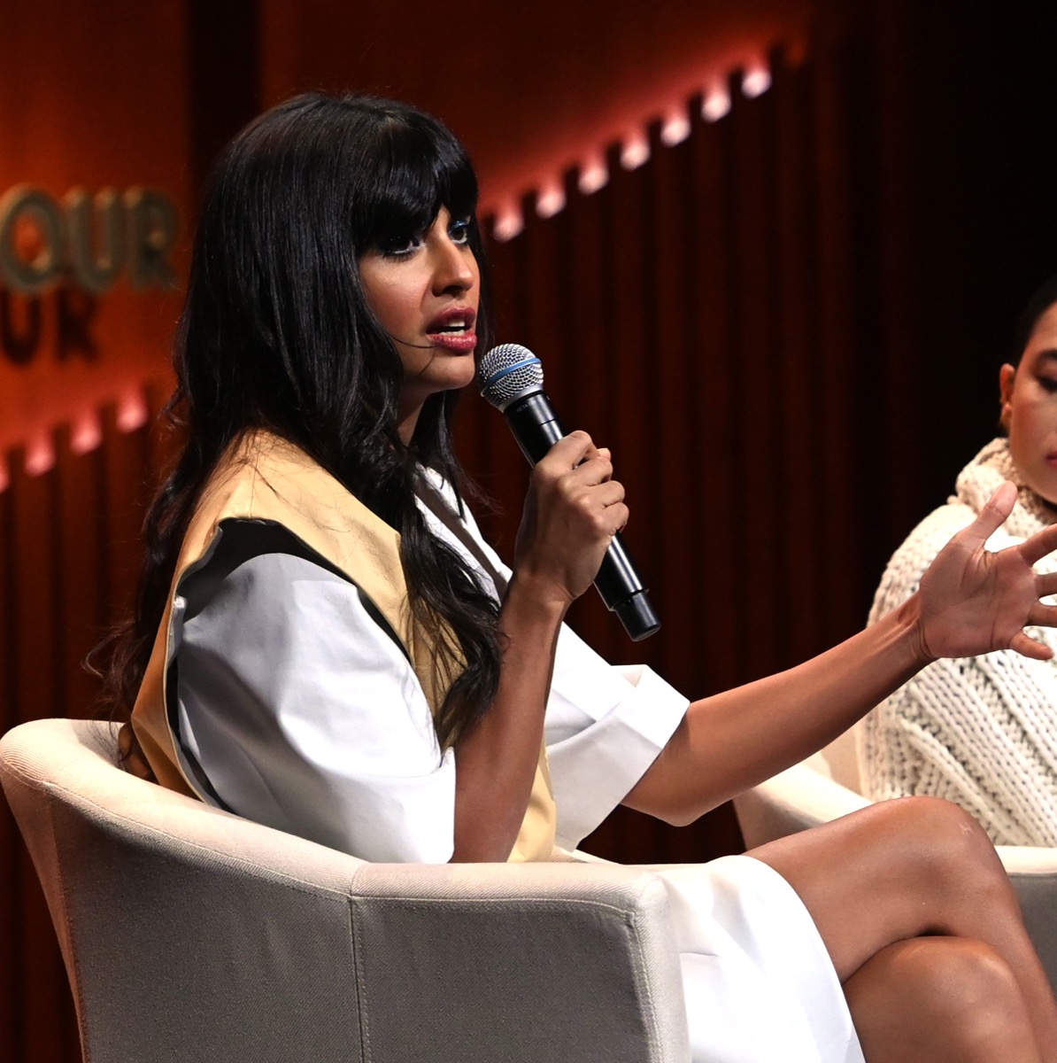 jameela jamil in white top and tan vest holding microphone while seated