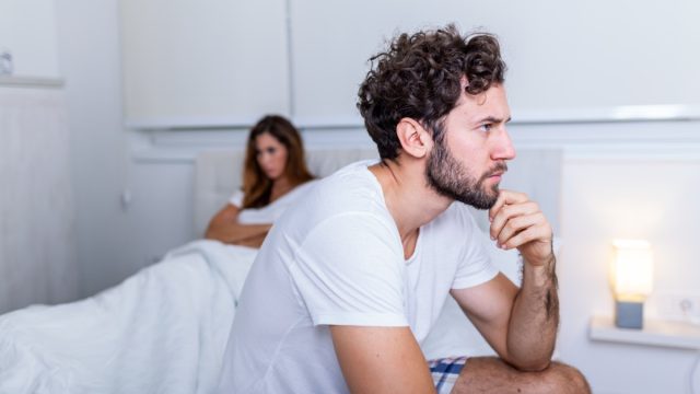 Sad man sitting on a bed, girlfriend in the background.