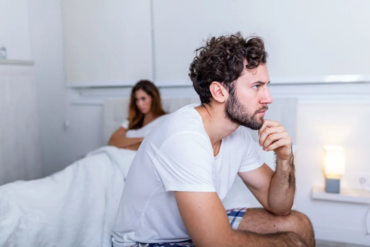 Hombre triste sentado en una cama, novia en el fondo.