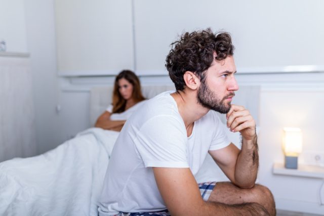 Sad man sitting on bed, girlfriend in background.