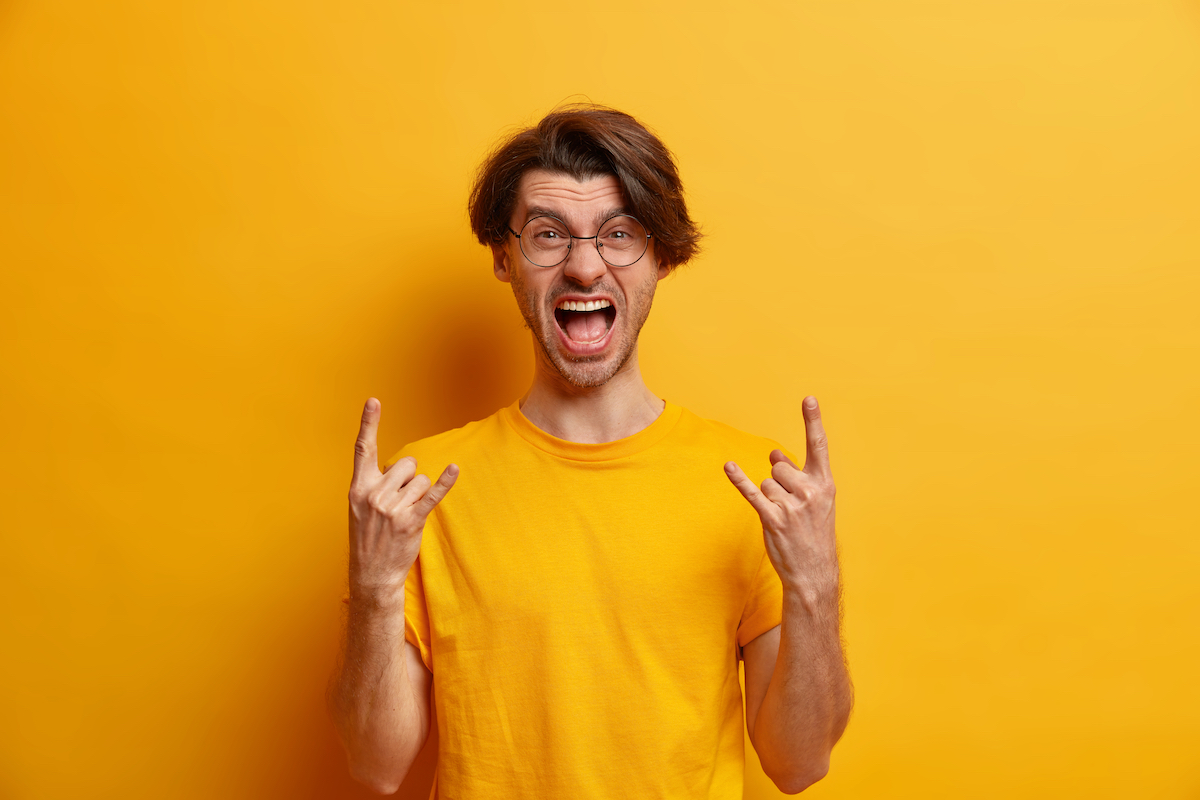 Rebelious unshaven hipster guy makes rock gesture screams out loudly being part of concert band wears round glasses and casual t shirt isolated over yellow studio background. Body language concept
