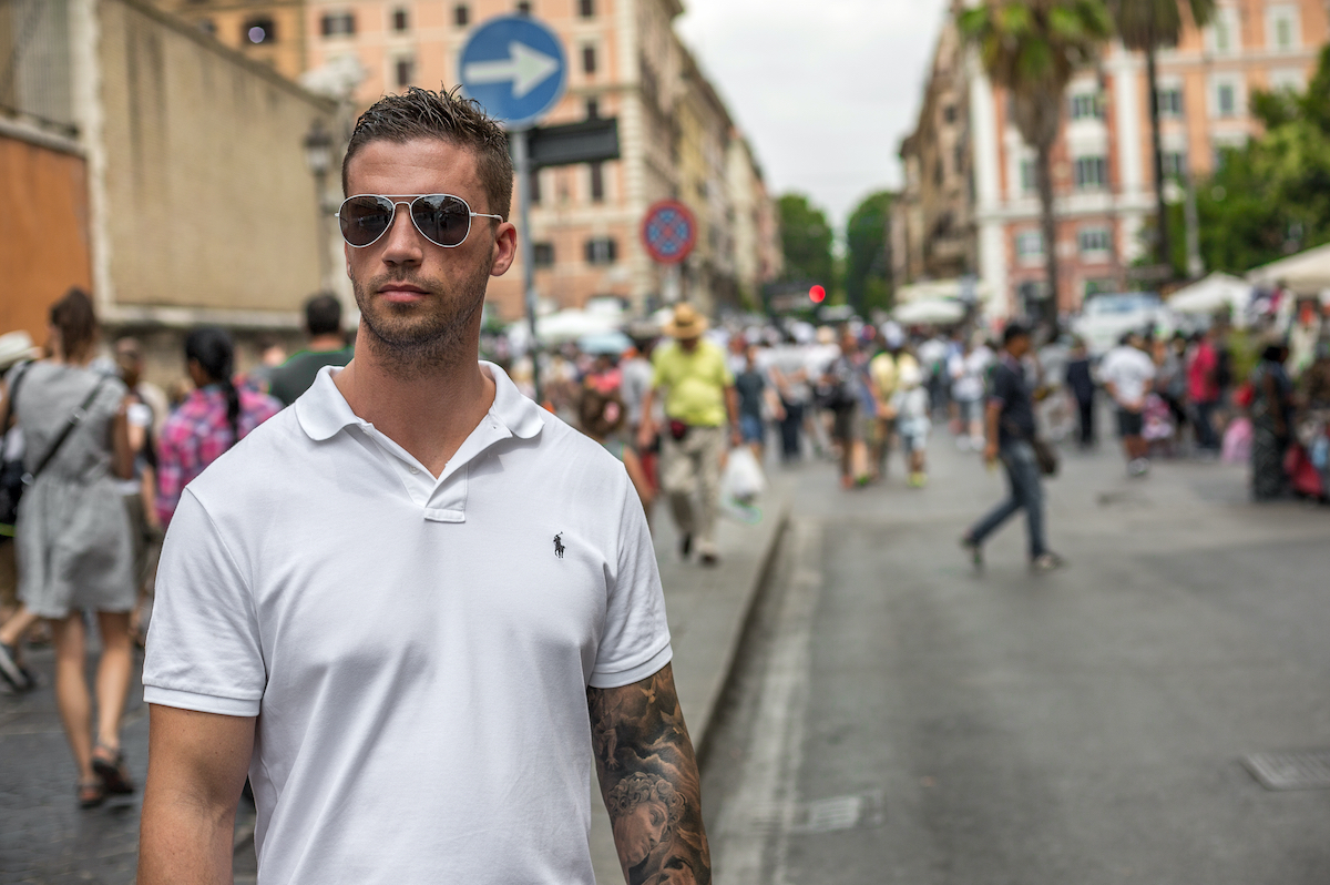 Rome, Italy - July 13th 2015: Cool handsome young caucasian man wearing sunglasses and a white t-shirt standing with confidence and style in a busy street as people walk past on the road and sidewalk