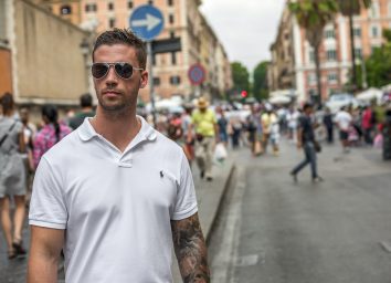 Rome, Italy - July 13th 2015: Cool handsome young caucasian man wearing sunglasses and a white t-shirt standing with confidence and style in a busy street as people walk past on the road and sidewalk